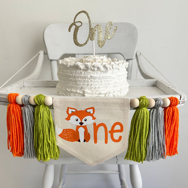 A 5 by 7 inch canvas flag with a cute fox followed by the letters NE in orange to spell the word ONE. On each side there is a 5 inch lime green, gray and orange yarn tassel each separated by a wooden bead for a total of 6 tassels and 6 beads.