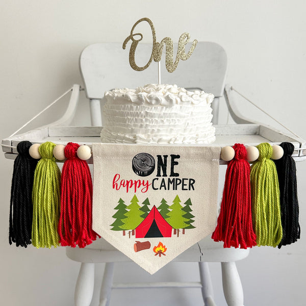 A 5 by 7 inch canvas banner with a log slice followed by the letters NE in black, with HAPPY CAMPER under the word ONE. There are images of a tent, trees and a campfire under the words. On each side is a red, lime green and black tassel with 6 beads.