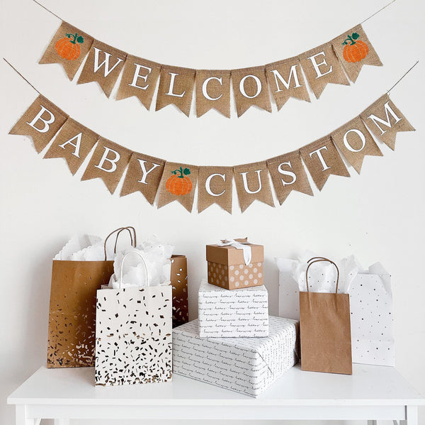 A burlap banner made from 4.5 by 6.5 inch fishtail shaped flags. The top string says WELCOME in white letters and has an orange pumpkin on each end. The bottom string says BABY followed by your choice of custom wording with a pumpkin in the middle.