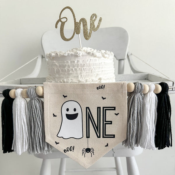 A 5 x 7 inch canvas center flag with a ghost followed by the letters NE in black and accented with a spider, bats and the word BOO. On each side there is a gray, white and black 5 inch yarn tassel, each separated by a wooden bead.