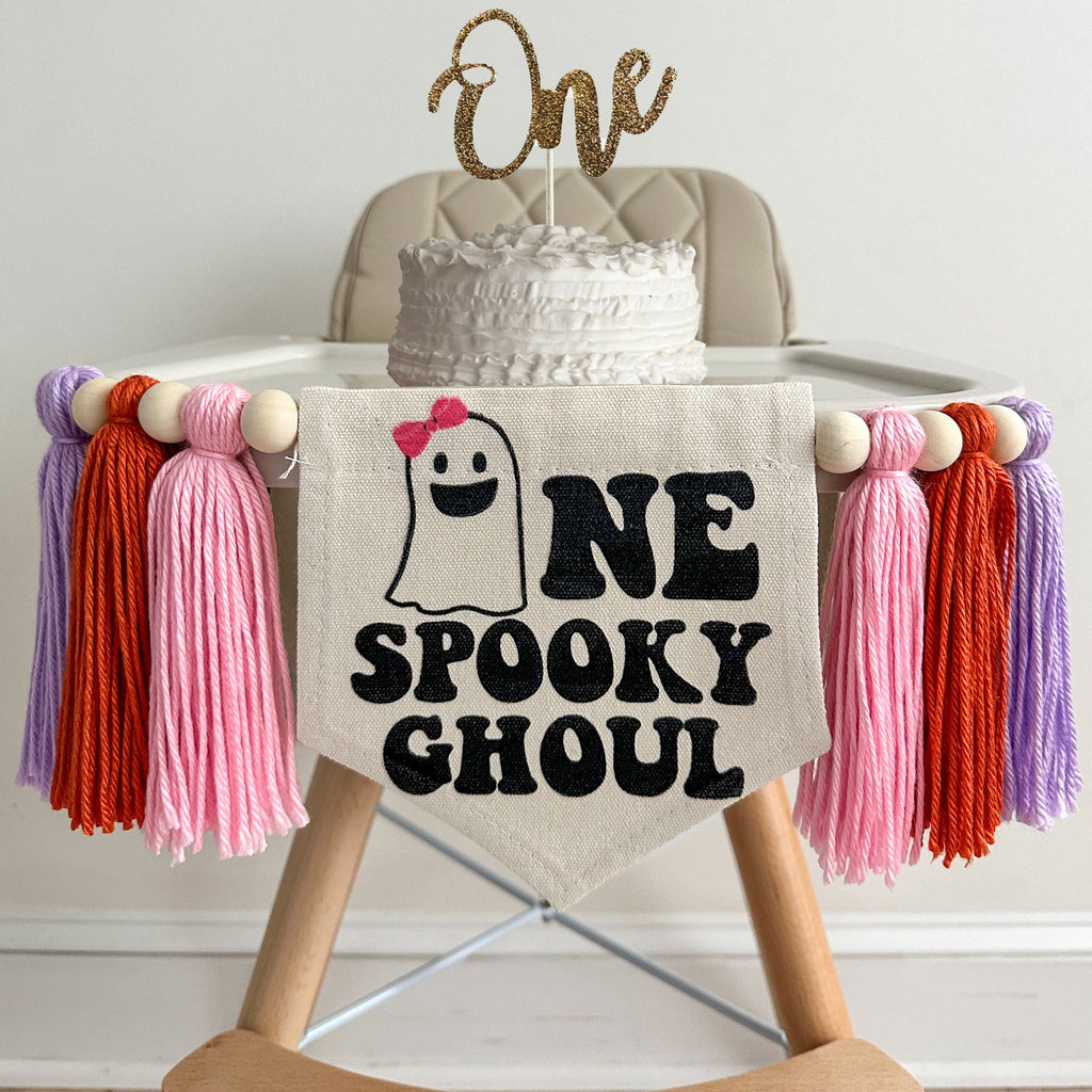 A 5 x 7 inch canvas center flag with a ghost with a pink bow followed by the letters NE in black and the word SPOOKY GHOUL underneath. On each side there is a pink, dark orange and lavender 5 inch yarn tassel, each separated by a wooden bead.