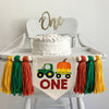 A 5 x 7 inch canvas center flag with a green tractor pulling a big orange pumpkin in a wagon. The word ONE is under it in reddish brown letters. On each side there is a dark orange, green and gold 5 inch yarn tassel, each separated by a wooden bead.