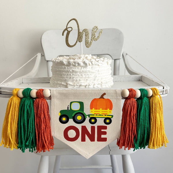 A 5 x 7 inch canvas center flag with a green tractor pulling a big orange pumpkin in a wagon. The word ONE is under it in reddish brown letters. On each side there is a dark orange, green and gold 5 inch yarn tassel, each separated by a wooden bead.