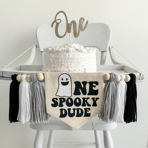 A 5 x 7 inch canvas center flag with a ghost followed by the letters NE in black and the words SPOOKY DUDE underneath. On each side there is a gray, white and black 5 inch yarn tassel, each separated by a wooden bead.