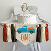 A 5 x 7 inch canvas center flag with blue truck full of pumpkins and the word ONE in orange under the truck. On each side there is a taupe brown, cream and dark orange 5 inch yarn tassel, each separated by a wooden bead.