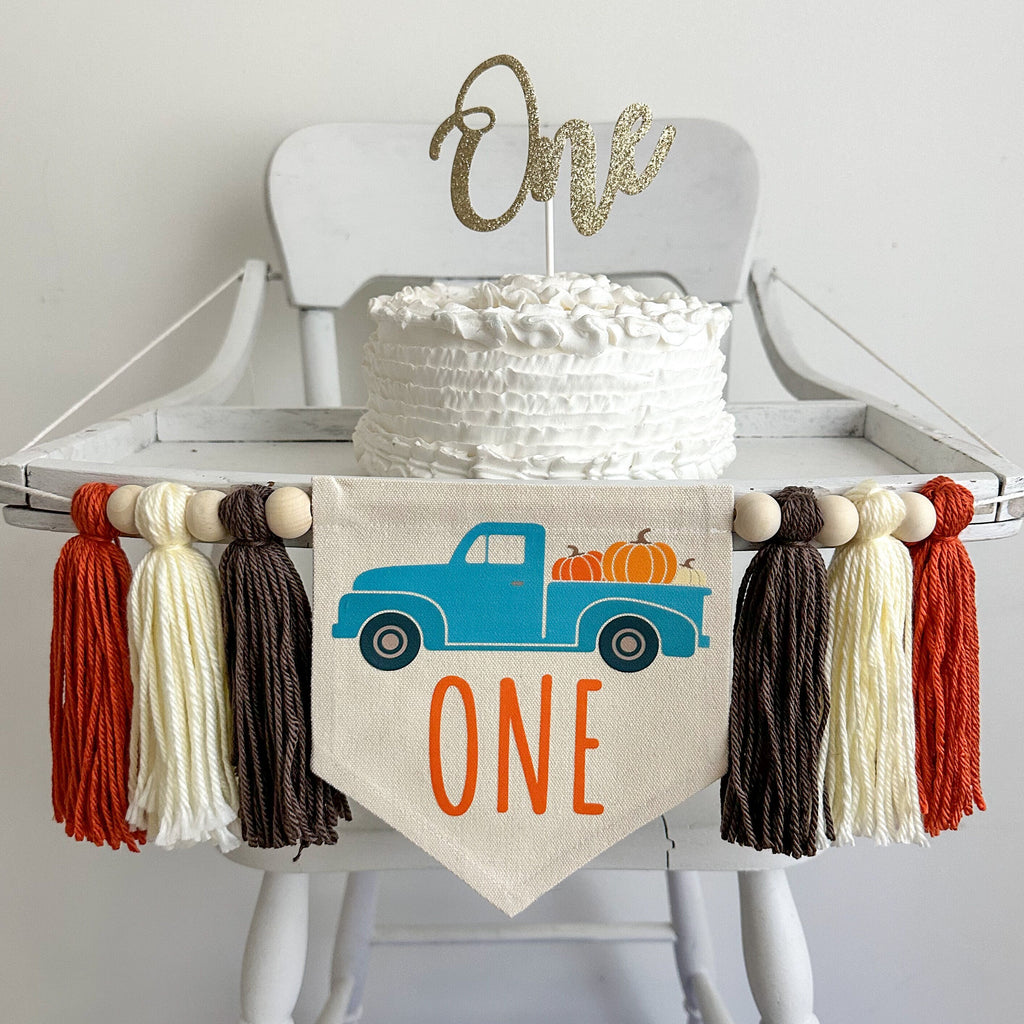 A 5 x 7 inch canvas center flag with blue truck full of pumpkins and the word ONE in orange under the truck. On each side there is a taupe brown, cream and dark orange 5 inch yarn tassel, each separated by a wooden bead.