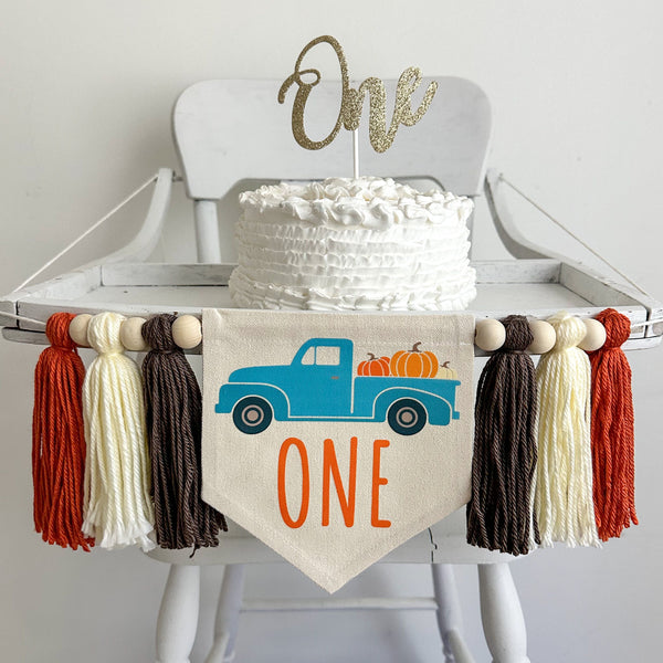 A 5 x 7 inch canvas center flag with blue truck full of pumpkins and the word ONE in orange under the truck. On each side there is a taupe brown, cream and dark orange 5 inch yarn tassel, each separated by a wooden bead.