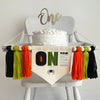 A 5 x 7 inch canvas center flag with the word ONE made from creepy Halloween characters like a spooky eye, mummy and spider. On each side there is an orange, lime green and black 5 inch yarn tassel, each separated by a wooden bead.