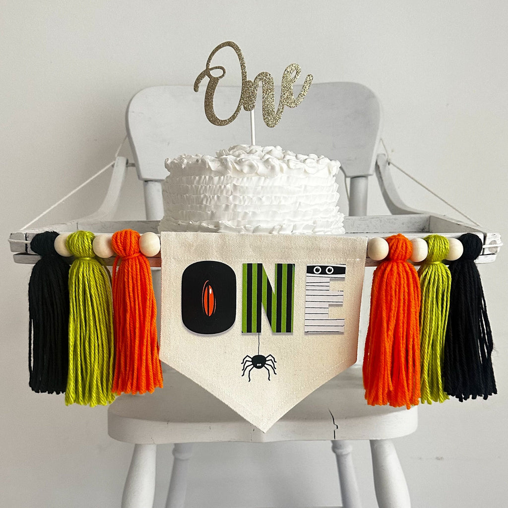 A 5 x 7 inch canvas center flag with the word ONE made from creepy Halloween characters like a spooky eye, mummy and spider. On each side there is an orange, lime green and black 5 inch yarn tassel, each separated by a wooden bead.