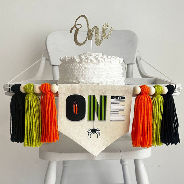 A 5 x 7 inch canvas center flag with the word ONE made from creepy Halloween characters like a spooky eye, mummy and spider. On each side there is an orange, lime green and black 5 inch yarn tassel, each separated by a wooden bead.