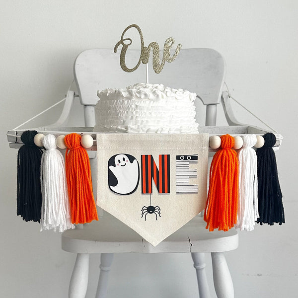 A 5 x 7 inch canvas center flag with the word ONE made from spooky characters like a ghost, spider and mummy. On each side there is an orange, white and black 5 inch yarn tassel, each separated by a wooden bead.