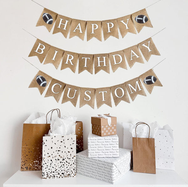 A burlap banner made from 4.5 by 6.5 inch fishtail shaped flags. The top line says HAPPY in white letters with a football on each end, the middle line says BIRTHDAY in white letters and the bottom line is customizable with a football on each end.
