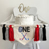 A 5 x 7 inch canvas banner with a hockey puck as the letter &quot;O&quot; followed by the letters NE in black. Under that is a pair of red and blue crossed hockey sticks. On each side is a red, gray and black 5 inch yarn tassel each separated by a wooden bead.