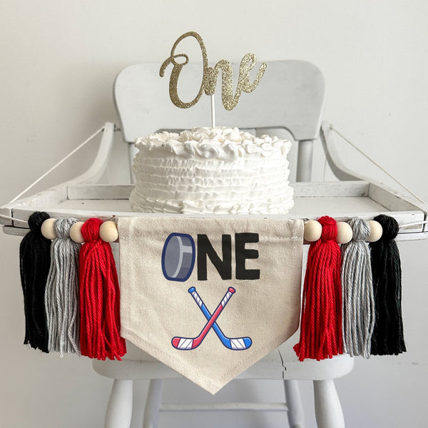 A 5 x 7 inch canvas banner with a hockey puck as the letter &quot;O&quot; followed by the letters NE in black. Under that is a pair of red and blue crossed hockey sticks. On each side is a red, gray and black 5 inch yarn tassel each separated by a wooden bead.