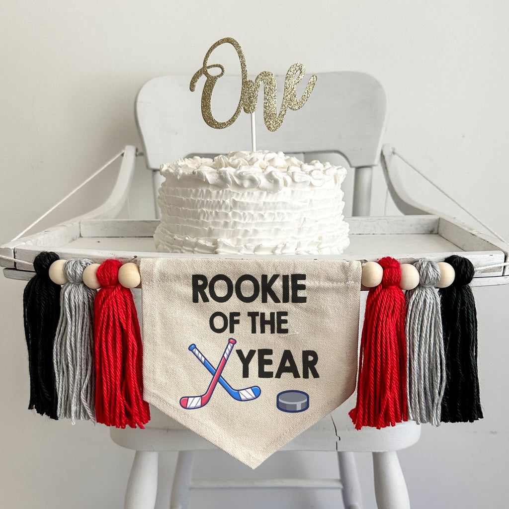 A 5 x 7 inch canvas banner with the words ROOKIE OF THE YEAR in black. Below the words there is a pair of crossed hockey sticks and a puck. On each side of the flag is a red, gray and black 5 inch yarn tassel each separated by a wooden bead.