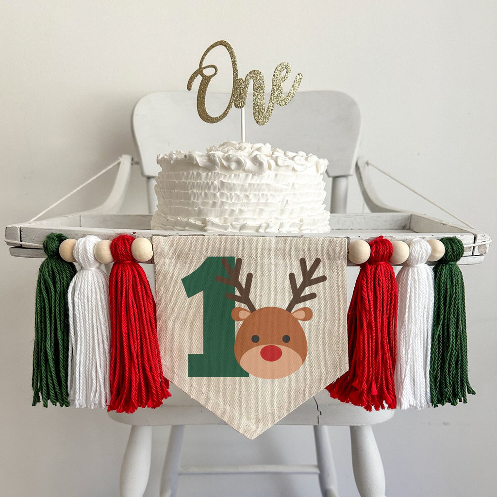 A 5 x 7 canvas center flag with a big green number 1 with a cute reindeer head beside it to the right. There is a red, white and dark green 5 inch yarn tassel on each side, each separated by a wooden bead for a total of 6 tassels and 6 beads.