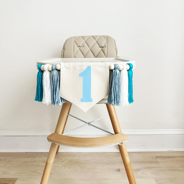 A 5 x 7 inch canvas flag with a light blue number 1 on it. On each side is a light blue, white and aqua colored 5 inch yarn tassel each separated by a wooden bead. There is a total of 6 tassels and 6 wooden beads.