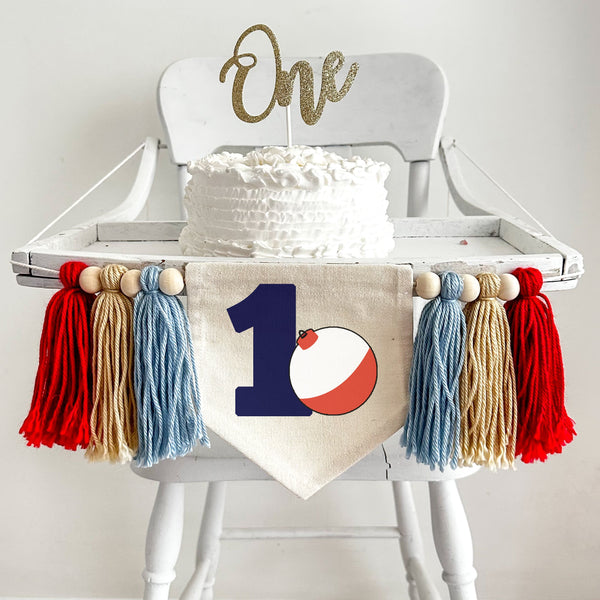 A 5 by 7 inch canvas banner with a large navy blue number 1 with a red and white fishing bobber propped beside the one. On each side there is a 5 inch light blue, tan and red yarn tassel, each separated by a wooden bead for a total of 6 tassels.