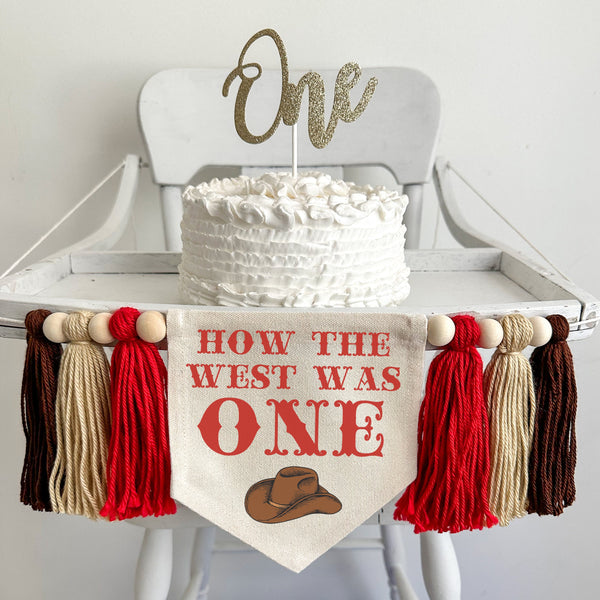 A 5 by 7 inch canvas flag with the words HOW THE WEST WAS ONE in red western style font with a brown cowboy hat under the words. On each side is a 5 inch tassel in red, tan and brown yarn each separated by a wooden bead. There are a total of 6 tassel
