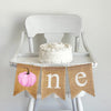 A burlap banner made from three 5 x 7 inch fishtail shaped flags. The first flag has a pink watercolor style pumpkin on it to create the letter O. The second flag has a lowercase letter N in white and the third flag has a lowercase letter E in white.