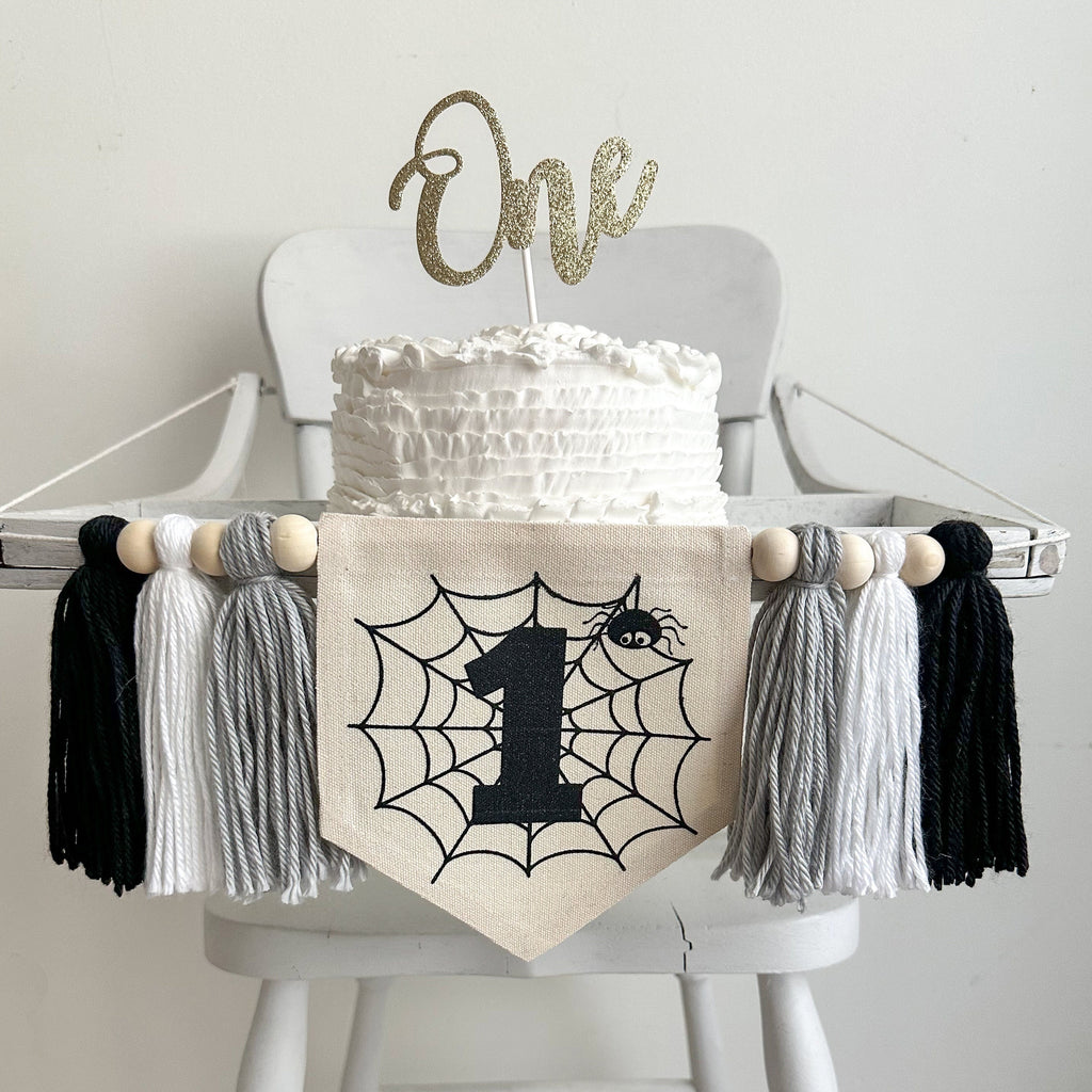 A 5 by 7 canvas flag with a spider web, black number 1 and a spider. On each side of the flag is a 5 inch yarn tassel in gray, white and black. Each tassel is separated by a wooden bead.