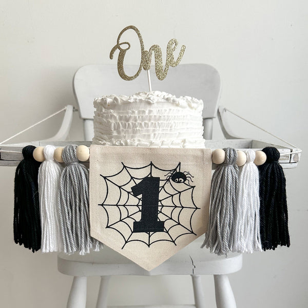 A 5 by 7 canvas flag with a spider web, black number 1 and a spider. On each side of the flag is a 5 inch yarn tassel in gray, white and black. Each tassel is separated by a wooden bead.