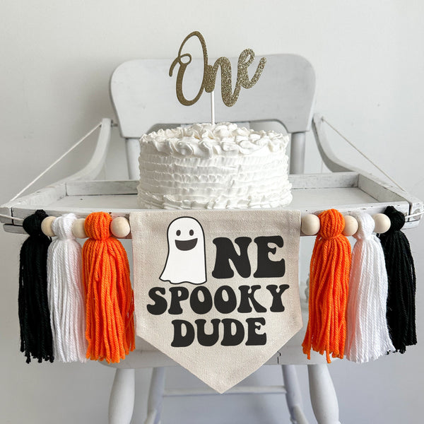 A 5 x 7 inch canvas center flag with a ghost followed by the letters NE in black. Under that is says SPOOKY ONE in black. There is an orange, white and black 5 inch yarn tassel on each side. They are each separated by a wooden bead for a total of 6.