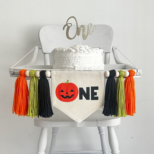 A 5 x 7 inch canvas center flag with an orange Jack O Lantern followed by the letters NE in black. On each side there is a black, lime green and orange 5 inch yarn tassel, each separated by a wooden bead.