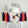 A 5 x 7 inch canvas center flag with the word ONE made from Halloween characters like a ghost, mummy and witch hat with a spider on the bottom. On each side there is a purple, orange and black 5 inch yarn tassel, each separated by a wooden bead.