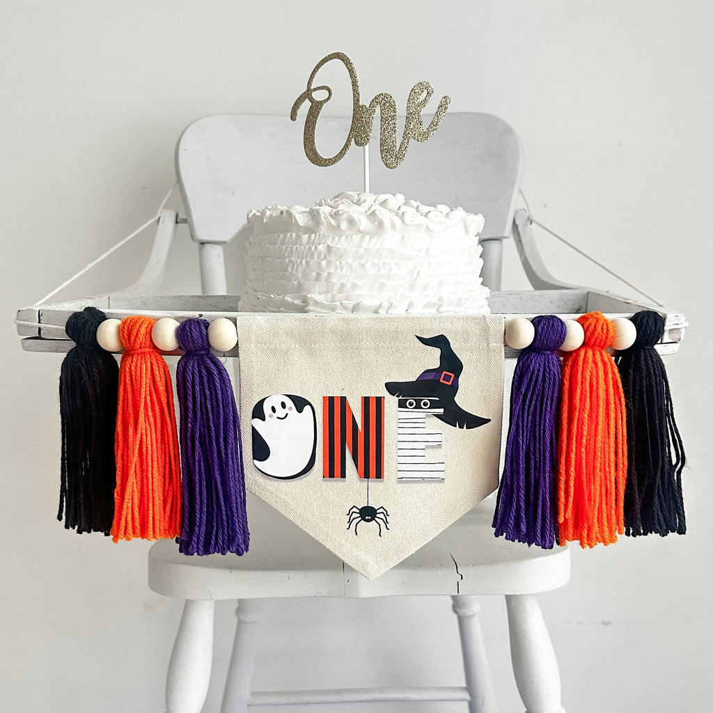 A 5 x 7 inch canvas center flag with the word ONE made from Halloween characters like a ghost, mummy and witch hat with a spider on the bottom. On each side there is a purple, orange and black 5 inch yarn tassel, each separated by a wooden bead.