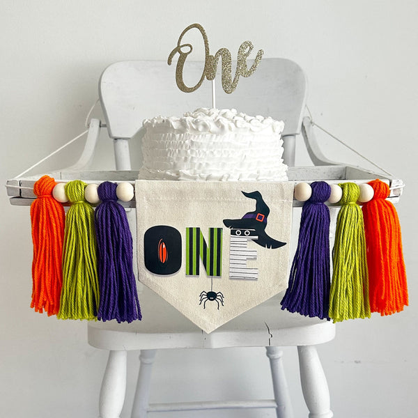 A 5 x 7 inch canvas center flag with the word ONE made from Halloween characters like a creepy eye, mummy, witch hat and spider. On each side there is a purple, lime green and orange 5 inch yarn tassel, each separated by a wooden bead.