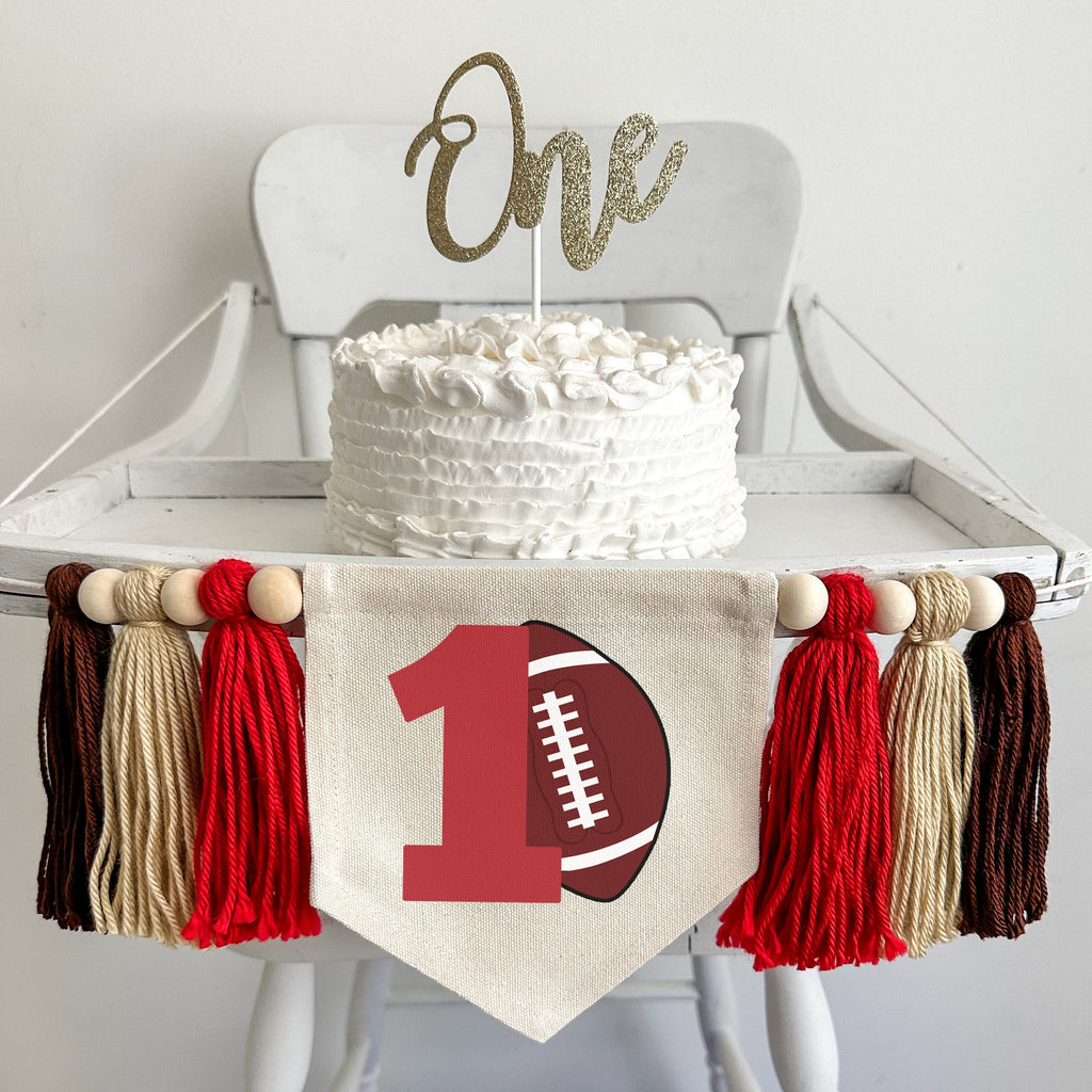A 5 x 7 canvas flag with a large red number 1 with a brown football propped against it. On each side there is a red, tan and brown 5 inch yarn tassel, each separated by a wooden bead for a total of 6 tassels and 6 beads.
