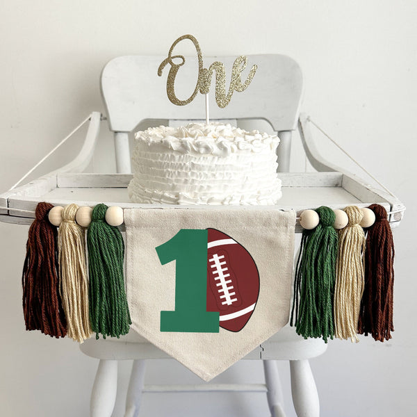 A 5 x 7 canvas flag with a large green number 1 with a brown football propped against it. On each side there is a dark green, tan and brown 5 inch yarn tassel, each separated by a wooden bead for a total of 6 tassels and 6 beads.