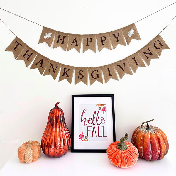 Happy Thanksgiving burlap banner. This banner has 2 lines, in brown letters the top layer spells out Happy with a white oak leaf on either side. The second line says Thanksgiving in brown letters.
