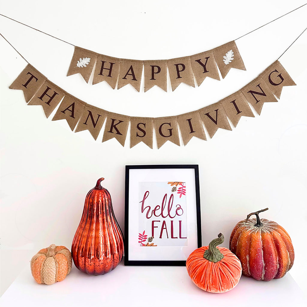 Happy Thanksgiving burlap banner. This banner has 2 lines, in brown letters the top layer spells out Happy with a white oak leaf on either side. The second line says Thanksgiving in brown letters.