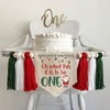 A 5 x 7 canvas center flag with the words OH WHAT FUN IT IS TO BE in red and the word ONE in green. There is a Santa on the right side and ornaments across the top. There is a red, white and green 5 inch tassel on each side with wooden beads by each.