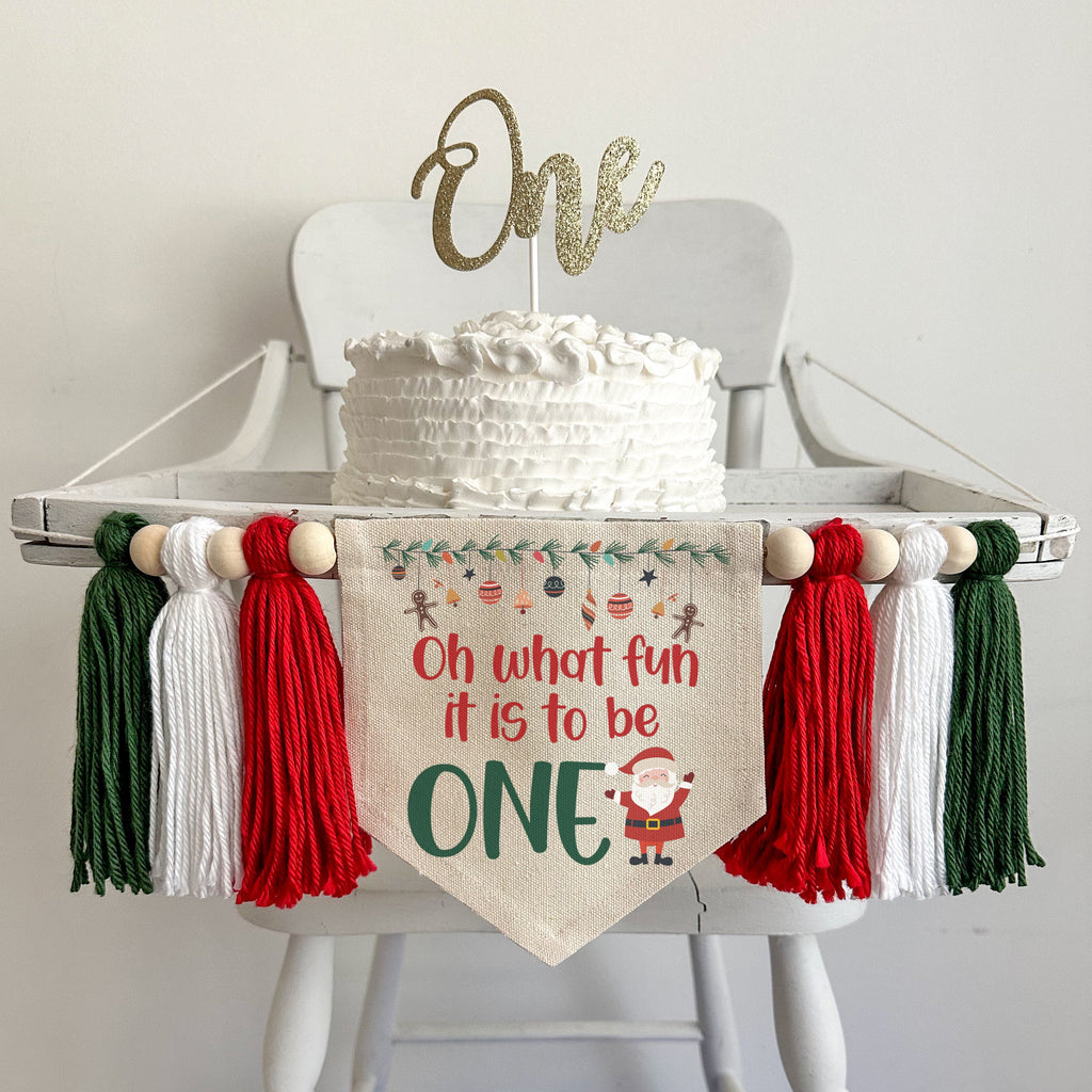 A 5 x 7 canvas center flag with the words OH WHAT FUN IT IS TO BE in red and the word ONE in green. There is a Santa on the right side and ornaments across the top. There is a red, white and green 5 inch tassel on each side with wooden beads by each.