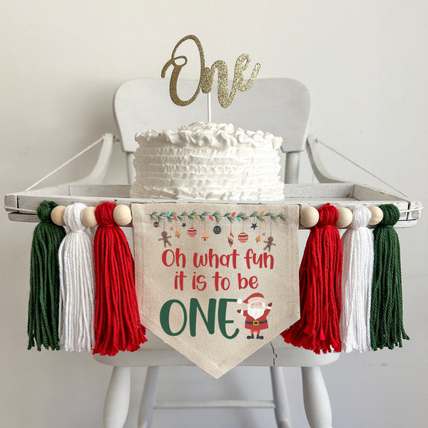 A 5 x 7 canvas center flag with the words OH WHAT FUN IT IS TO BE in red and the word ONE in green. There is a Santa on the right side and ornaments across the top. There is a red, white and green 5 inch tassel on each side with wooden beads by each.
