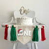 A 5 x 7 canvas center flag with the word ONE in white with a shadow behind it. There is a Santa hat sitting on top of the O. There is a green, white and red 5 inch yarn tassel on each side, each separated by a wooden bead.
