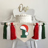 A 5 x 7 canvas center flag with a dark green number 1 with a Santa hat sitting on top of it. On each side there is a red, white and dark green 5 inch yarn tassel, each separated by a wooden bead for a total of 6 tassels and 6 beads.