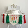A 5 x 7 canvas center flag with the words OH WHAT FUN TO BE in red and the word ONE in green. There is a gingerbread man on the left side. There is a green, white and red 5 inch yarn tassel on each side, each separated by a wooden bead.