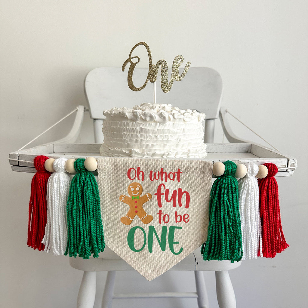 A 5 x 7 canvas center flag with the words OH WHAT FUN TO BE in red and the word ONE in green. There is a gingerbread man on the left side. There is a green, white and red 5 inch yarn tassel on each side, each separated by a wooden bead.