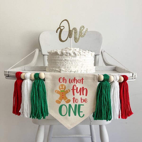 A 5 x 7 canvas center flag with the words OH WHAT FUN TO BE in red and the word ONE in green. There is a gingerbread man on the left side. There is a green, white and red 5 inch yarn tassel on each side, each separated by a wooden bead.