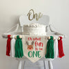 A 5 x 7 canvas center flag with the words OH WHAT FUN TO BE in red and the word ONE in green. There is a reindeer on the left side. There is a green, white and red 5 inch yarn tassel on each side, each separated by a wooden bead.