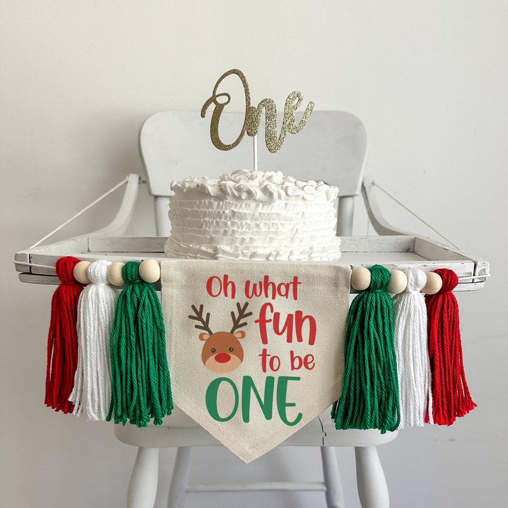 A 5 x 7 canvas center flag with the words OH WHAT FUN TO BE in red and the word ONE in green. There is a reindeer on the left side. There is a green, white and red 5 inch yarn tassel on each side, each separated by a wooden bead.
