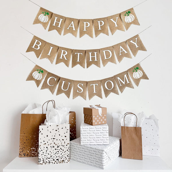 A burlap banner made from 4.5 by 6.5 inch fishtail shaped flags in white letters. The top line says HAPPY with a white pumpkin on each end. The middle line says BIRTHDAY and the bottom line can be custom text of your choice with a pumpkin on each end