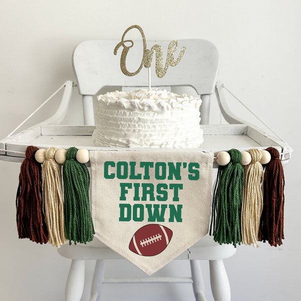 A 5 by 7 inch canvas flag with a custom name followed by the words FIRST DOWN in green. There is a brown football under the words. On each side is a dark green, tan and brown 5 inch yarn tassel, each separated by a wooden bead, six tassels in total.