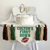 A 5 by 7 inch canvas flag with a custom name followed by the words FIRST DOWN in green. There is a brown football under the words. On each side is a dark green, tan and brown 5 inch yarn tassel, each separated by a wooden bead, six tassels in total.