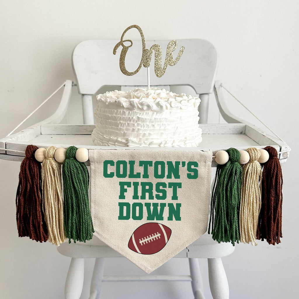 A 5 by 7 inch canvas flag with a custom name followed by the words FIRST DOWN in green. There is a brown football under the words. On each side is a dark green, tan and brown 5 inch yarn tassel, each separated by a wooden bead, six tassels in total.