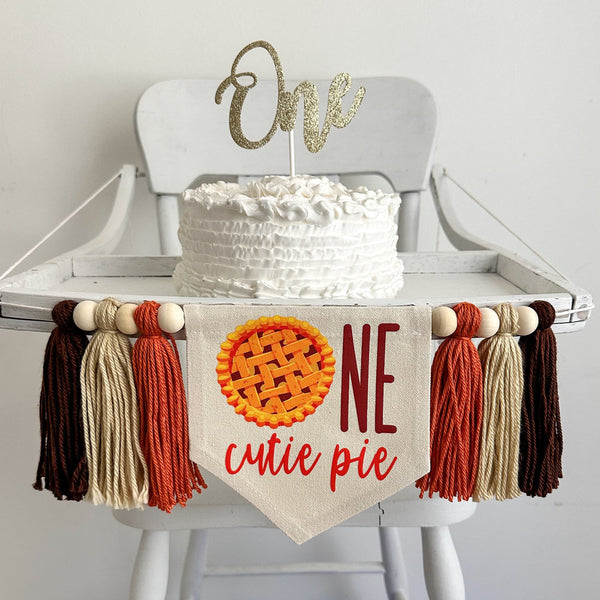 A 5 by 7 inch canvas banner with an apple pie image as an O followed by the letters NE in brown to spell the word ONE. The words CUTIE PIE are under in dark orange. On each side there is a dark orange, tan and brown tassel separated by wooden beads.